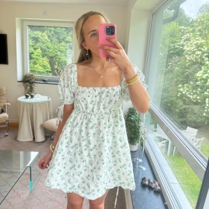 Vit blommig kortklänning med puffärm - Supersöt vit kortklänning med grönt blommönster, puffärmar med knyt och fyrkantig urringning. Klänningen har en flowy passform och känns perfekt för varma dagar. Materialet är lätt och andas, vilket gör den extra skön att bära. Fickor finns