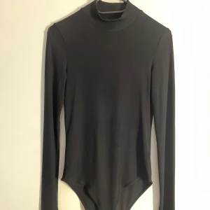 Svart ribbad polotopp från H&M - Svart långärmad polotopp från H&M i ribbat material. Kroppsnära passform med hög hals och tryckknappar i grenen. Perfekt att styla med jeans eller kjol för en clean look.