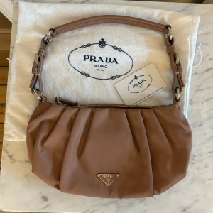 Beige axelväska från Prada med detaljer - Säljer en beige axelväska från Prada med coola broderade och metalliska detaljer på ena sidan. Väskan har ett mjukt, veckat tyg och justerbar axelrem i matchande färg. Klassisk Prada-logga framtill och dragkedja upptill.