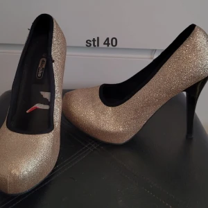 Glittriga pumps med hög klack - Snygga pumps i guldigt glittrigt material med svarta detaljer runt öppningen. Skorna har hög klack och rund tå, perfekta för att sticka ut och ge extra glam till din outfit.