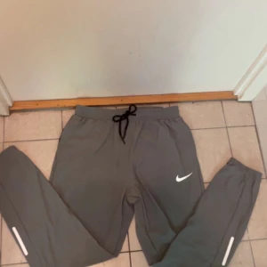 Grå träningsbyxor från Nike - Snygga grå träningsbyxor från Nike med svart snörning i midjan och vita reflexdetaljer på benen. Byxorna har en smal passform och är perfekta för löpning eller gym. Tillverkade i ett lätt och syntetiskt material som andas.