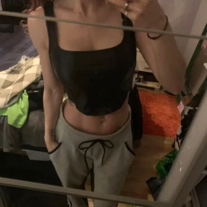 Svart korsett-topp i skinnimitation - Säljer en svart korsett-topp med breda axelband och markerade sömmar framtill. Toppen har en croppad passform och är gjord i skinnimitation som ger en edgy look. Perfekt att styla med jeans eller kjol för en cool outfit.