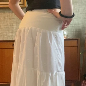 Vit långkjol med broderade blommor - Superfin vit långkjol med volanger som är i 100% lätt bomullstyg och är uppsydd i en ateljé. Kjolen har bred resår i midjan, och är halvvägs fodrad så att kjolen inte är genomskinlig. Midjan rakt över är 33cm och kjolens längd är 88cm, själv är jag ungefär 160cm lång. Perfekt sommar kjol. 