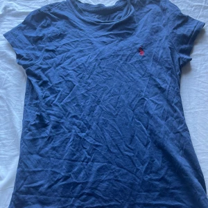 Blå Ralph lauren t-shirt  - Klassisk blå t-shirt från Polo Ralph Lauren med röd broderad logga på bröstet. T-shirten har rund halsringning och korta ärmar. Tillverkad i mjuk bomull som känns skön mot huden. Perfekt för en avslappnad och stilren look.