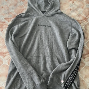 Grå hoodie från Champion XL - Snygg grå hoodie från Champion i storlek XL. Tröjan har svart Champion-logga på bröstet och svarta band med logga längs ärmarna. Klassisk känguruficka och huva. Mjuk bomullsblandning som är perfekt för chill eller streetwear.
