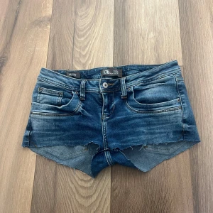 Blå jeansshorts från LTB - Snygga blå jeansshorts från LTB med råa, fransiga kanter och klassisk femficksdesign. Shortsen har låg midja och slitna detaljer för en cool look. Perfekta för sommardagar och festivaler. Har gjort små hål på insidan för dra igen ett skosnöre men inget som syns eller märks. Midjan storlek 27