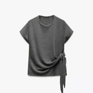 Grå topp med knyt från Zara - Stilren grå topp från Zara med korta ärmar och rund halsringning. Toppen har en snygg knytdetalj i sidan som ger en cool twist till looken. Perfekt att matcha med jeans eller kjol för en enkel men trendig outfit.