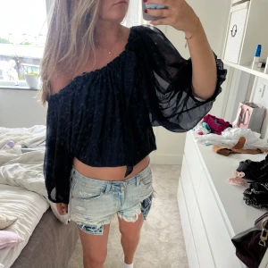 Svart offshoulder blus med volymärmar - Säljer en svart, luftig offshoulder blus med transparenta, voluminösa långärmar. Toppen har en rynkad resårkant upptill som ger en snygg passform över axlarna. Perfekt för dig som gillar trendiga och unika plagg.