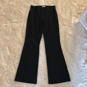 Svarta bootcut kostymbyxor från Nelly - Svarta kostymbyxor från Nelly i bootcut-modell. Byxorna har klassiska fickor fram, hällor för bälte och är tillverkade i ett mjukt tyg som ger en snygg siluett. Perfekta för en clean och stilren look.