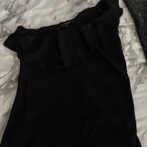 Svart oneshoulder topp från KappAhl - Snygg svart oneshoulder topp från KappAhl med volangdetalj längs halsringningen. Perfekt för dig som vill ha en stilren och trendig look. Tunn och lätt i materialet, passar till många olika outfits.