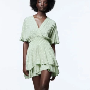 Grön prickig kortklänning från Zara - Supersöt ljusgrön kortklänning från Zara med vita prickar, v-ringning och smockad midja. Klänningen har volangdetaljer i kjolen och ärmarna, samt korta ärmar med liten volangkant. Perfekt för sommarens alla tillfällen! 