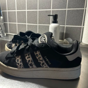 Adidas Campus sneakers med leopardmönster - Svarta Adidas Campus sneakers med klassiska tre ränder i beige leopardmönster. Skorna har grova svarta snören, vit sula och detaljer i mocka. Adidas-logga på plös och häl. Perfekta för dig som vill sticka ut med en cool twist på en ikonisk modell.