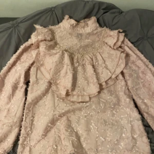 spetsblus med volang och pärldetaljer - Supersöt puderrosa blus med struktur, volanger och pärldetaljer framtill. Hög krage och knappar i hals och ärmslut. Perfekt för både vardag och fest! Använd få gånger 