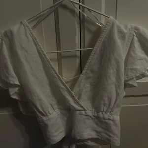 Vit omlottopp från Bik Bok XS - Supersöt vit omlottopp från Bik Bok i storlek XS. Toppen har korta volangärmar, djup v-ringning och knyt i midjan. Tillverkad i ett luftigt linnematerial, perfekt för varma dagar. Enkel att styla med både jeans och kjol.