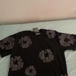 Svart t-shirt från Denim Tears - Svart t-shirt från Denim Tears med unikt blommigt mönster i grått över hela tröjan. Klassisk passform och rund halsringning. Perfekt för dig som vill sticka ut med en cool och modern stil. Priset kan diskuteras.
