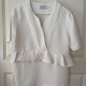 Vit blus med volang från & Other Stories - Supersöt vit blus från & Other Stories med v-ringning och volangdetalj längs midjan. Blusen har korta ärmar och är gjord i ett mjukt, lätt material som ger en lyxig känsla. Perfekt för dig som gillar stilrena och feminina plagg.