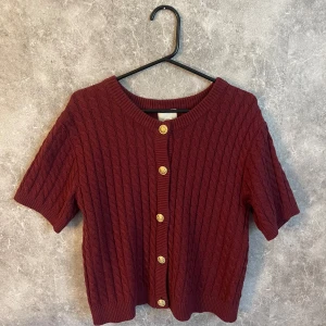 Vinröd kabelstickad kofta med guldknappar - Vinröd kabelstickad kofta med kort ärm och guldknappar framtill. Tröjan har rund halsringning och ribbade muddar. Koftan är som i nytt skick och har andvänts mycket fåtal gånger. Brukar vanligtvis ha S i storlek men L i denna eftersom den var liten i storlek.