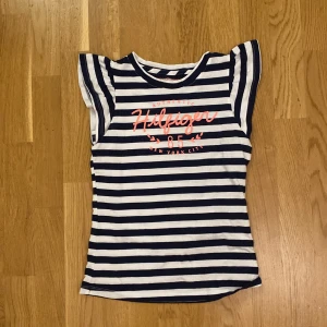 Topp från Tommy Hilfiger - Snygg randig topp från Tommy Hilfiger i marinblått och vitt med rosa tryck framtill. Modellen är ärmlös med volangliknande axlar och rund halsringning. Stlk 12/14 år motsvarande XS
