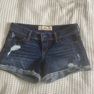 Mörkblå jeansshorts från Hollister - Snygga mörkblå jeansshorts från Hollister med slitna detaljer och fransig kant. Klassisk femficksmodell med orangea sömmar och uppvikta ben. Perfekta för sommaren och ger en avslappnad vibe.