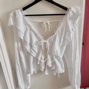 Vit blus  från Gina Tricot - Supersöt vit blus från Gina Tricot med volanger, knytning framtill och spetsdetaljer längs v-ringningen. Luftig och perfekt nu inför sommaren. Kommer med etikett💗