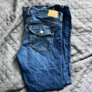 Blå jeans från Gina Tricot - Snygga blå jeans från Gina Tricot med klassisk femficksdesign och kontrastsömmar. Jeansen har en bakficka med lock och knappdetalj samt en läderpatch med logga i midjan. Perfekta för en avslappnad och trendig look.