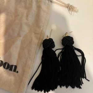 Svarta tofsörhängen med knut - Stilrena svarta örhängen med dekorativ knut upptill och långa tofsar som ger en cool och trendig look. Fästen i guldfärgad metall. Perfekta för att lyfta vilken outfit som helst.