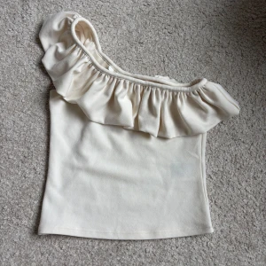 Offshoulder volangtopp i beige XS - Supersöt offshoulder topp i ljusbeige med bred volang längs ena axeln. Sitter tight och har en stilren, enkel design som passar perfekt till jeans eller kjol. Mjuk och skön känsla, perfekt för varma dagar. Jättesöt verkligen och aldrig använd🥰