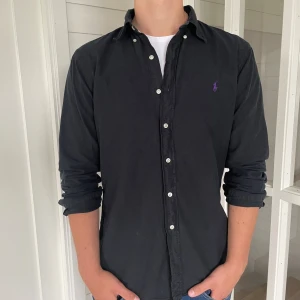 Ralph lauren skjorta - Säljer denna riktigt snygga svarta Ralph lauren skjorta med lila märke, den är i mycket bra skick utan några defekter, Storlek L modell slim fit den är 77 cm lång och 53 cm bred under armhålorna, hör gärna av dig vid frågor!