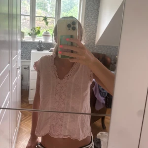 Ljusrosa blus med spetsdetaljer - Supersöt ljusrosa blus med broderade spetsdetaljer och små prickar i tyget. Blusen har korta ärmar med volangkant och v-ringning med dekorativ spets runt halsen. Luftig och flowy passform, perfekt för varma dagar.