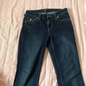 True Religion Becca Bootcut Jeans - Mörkblå jeans från True Religion, modell Becca Mid Rise Bootcut. Klassisk femficksdesign med snygga kontrastsömmar och ikonisk fickdetalj bak. Bootcut-ben och midwaist passform. Perfekta för dig som gillar en tidlös jeanslook med lite edge. Är inte helt säker då jag har köpt dom av min kompis men jag har för mig att dom är uppsydda nån centimeter.