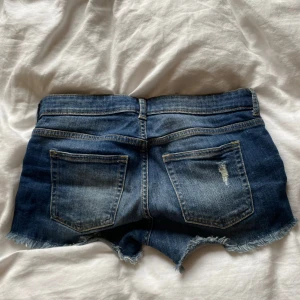 Blå jeansshorts med slitningar - Snygga blå jeansshorts med slitna detaljer och fransig kant nertill. Klassisk femficksmodell med låg midja och dragkedja. Perfekta för varma sommardagar och ger en avslappnad vibe🩷sparsamt använda! Skriv vid frågor🫶🏼