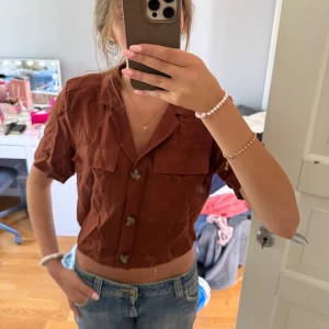 Brun croppad skjorta från Gina Tricot - Säljer en brun croppad blus från Gina Tricot med korta ärmar och stora knappar framtill. 