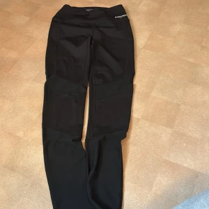 Svarta träningstights från Björn Borg - Svarta träningstights från Björn Borg med hög midja och tight passform. Tillverkade i stretchigt syntetmaterial som andas, med förstärkta partier på benen för extra hållbarhet. Perfekta för gymmet eller löprundan.