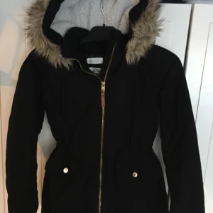 Svart parkajacka med fuskpäls från H&M - Svart parkajacka från H&M med mysig fuskpäls runt huvan och vit teddyfoder på insidan. Jackan har dragkedja framtill, två stora fickor med knappar och markerad midja. Perfekt för kalla dagar och riktigt snygg till vardagslooken.