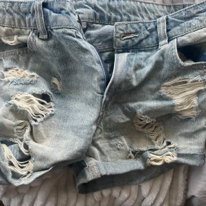 Ljusblå slitna jeansshorts - Snygga ljusblå jeansshorts med rejält slitna detaljer och fransiga hål framtill och baktill. Klassisk femficksmodell med uppvikta benslut och bälteshällor. Perfekta för en avslappnad och trendig sommarlook.