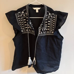 Svart broderad blus från H&M - Svart blus från H&M med vita broderier och snygga tofsar i snören framtill. Blusen har volangärmar och v-ringning, samt detaljerade broderier över axlar och bröst. Perfekt för dig som gillar boho-stil och unika detaljer.