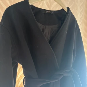 Svart omlottkappa från BikBok XS - Stilren svart kappa från BikBok i omlottmodell med bälte i midjan. Jackan har en klassisk rak passform, är helsvart och fodrad på insidan. Perfekt för dig som gillar minimalistisk och clean stil.