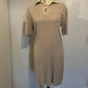 Beige ribbad kortklänning med krage - Ribbad beige kortklänning med v-ringning och klassisk krage. Klänningen har korta ärmar och en figurnära passform som ger en clean och stilren look. Perfekt för dig som gillar minimalistisk stil och mjuka material.