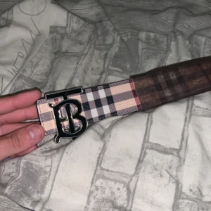 Burberry vändbart bälte med rutigt mönster - Snyggt vändbart bälte från Burberry med klassiskt rutigt mönster på ena sidan i beige, svart och rött, och helsvart med diskret mönster på andra sidan. Bältet har en stor metallspänne med Burberrys ikoniska B-logga. Perfekt accessoar för att lyfta din outfit.