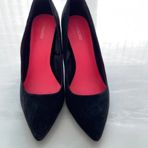 Svarta pumps från Even&Odd med klack - Stilrena svarta pumps från Even&Odd med spetsig tå och chunky hög klack. Skorna har en snygg röd innersula som ger en cool kontrast. Yttermaterialet är mocka-liknande och passar perfekt till dig som gillar en klassisk men edgy look. Använda endast en gång