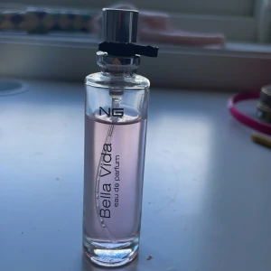 Bella Vida Eau de Parfum 15ml - Bella Vida Eau de Parfum från NG i en smal, cylindrisk glasflaska med silverfärgad spraykork. Parfymen har en ljusrosa ton och rymmer 15 ml. Flaskan har stilren svart text och en enkel, modern design som känns fräsch och ungdomlig. Mycket kvar! 