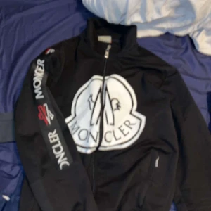 Svart Moncler jacka med stor logga - Svart jacka från Moncler med stor vit logga framtill och tryck på ena ärmen. Jackan har dragkedja och hög krage, perfekt för dig som gillar streetwear och vill sticka ut. Snygg och sportig look med tydliga Moncler-detaljer.