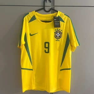 Brasilien Ronaldo 9 Nike fotbollströja - Gul Brasilien fotbollströja från Nike med gröna detaljer, nummer 9 och Ronaldo på ryggen. Klassisk CBF-logga på bröstet och Nike Swoosh. Tillverkad i lätt och ventilerande material, perfekt för match eller träning.