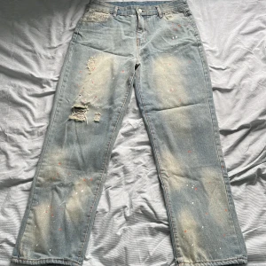 Ljusblå distressed jeans med färgstänk - Säljer ett par ljusblå jeans med raka ben, slitna detaljer och coola färgstänk i rött och vitt. Jeansen har klassisk femficksdesign och en avslappnad vibe. Perfekta för dig som gillar en unik och kreativ look.