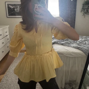 Gul blus med volang och knyt - Supersöt gul blus med volangkant nedtill och smockad överdel. Blusen har trekvartsärm med puff och knytning vid halsen för extra detalj. Perfekt för dig som vill ha en färgglad och luftig topp med lite romantisk känsla.
