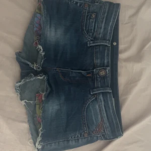 Blå jeansshorts från Replay - Snygga blå jeansshorts från Replay med slitna kanter och coola detaljer på bakfickorna. Klassisk femficksmodell med knapp och dragkedja framtill. Shortsen har en mörkblå tvätt och synliga slitningar för en avslappnad vibe. Perfekta för sommaren!