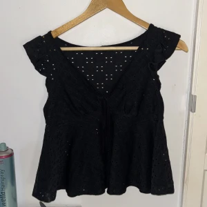 Svart blus med knyt och broderade hål - Supersöt svart blus med v-ringning, knyt framtill och broderade hål som ger en luftig känsla. Volangiga ärmar och lätt utsvängd passform. Perfekt för dig som gillar romantiska detaljer och en chill men ändå snygg look.