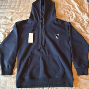 Marinblå hoodie från Ami Paris - Snygg marinblå hoodie från Ami Paris med klassisk känguruficka och broderad logga på bröstet. Tröjan har huva med snörning och ribbade muddar. Perfekt för en chill och stilren look.