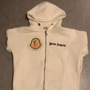Moncler x Palm Angels zip hoodie - Vit hoodie med dragkedja från Moncler x Palm Angels. Zip hoodien har, stor huva och tydliga loggor på bröstet. Materialet är mjukt. Snygg broderad Moncler-patch och Palm Angels-tryck framtill.