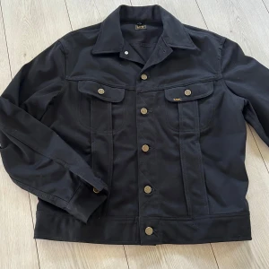 Svart jeansjacka från Lee Riders strl 52 - Klassisk svart jeansjacka från Lee Riders med snygga bronsfärgade knappar och två bröstfickor med lock. Jackan har en rak passform, krage och tydliga sömmar som ger en stilren look. Perfekt för dig som gillar tidlös streetstyle.  Jackan är helt ny och kommer från djur och rökfritt hem 